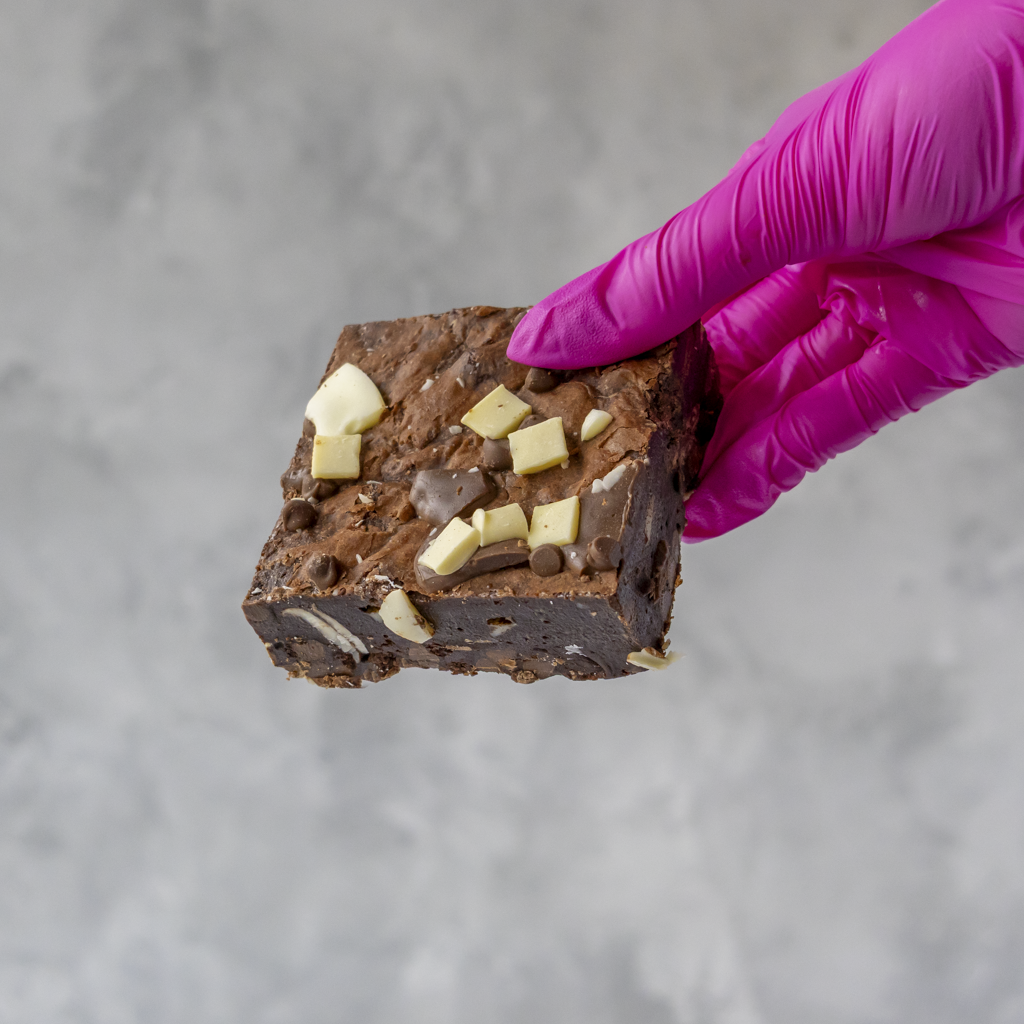 Brownie with chocolate chips and white pieces held by a hand in a pink glove on a gray background