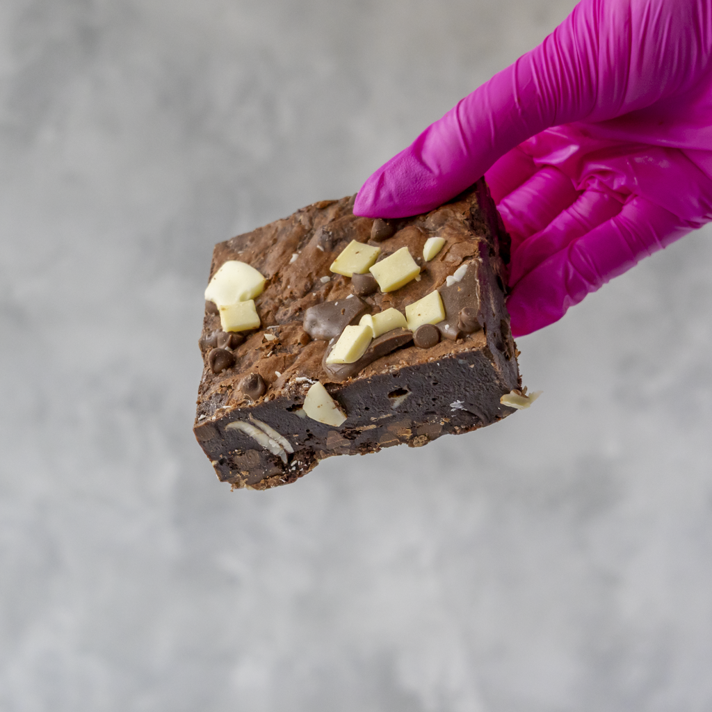 Chocolate brownie with white and milky chocolate chips held by a gloved hand on a gray background