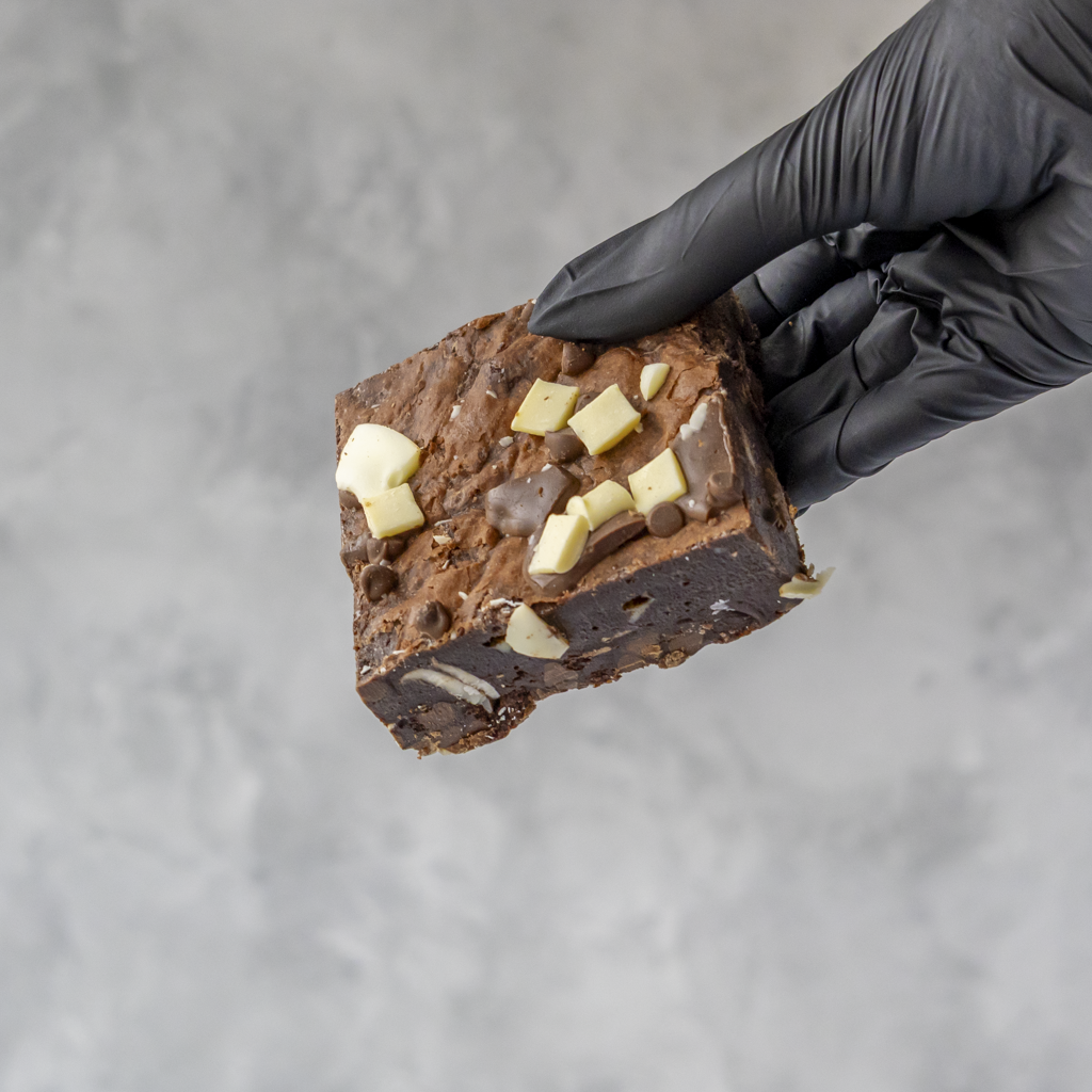 Hand wearing a black glove holding a piece of chocolate brownie with chocolate chips against a gray background