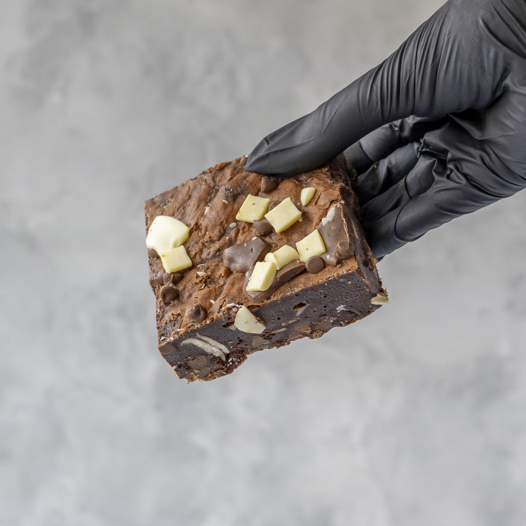 Chocolate brownie with milk, dark and white chocolate chips held by a gloved hand on a gray background