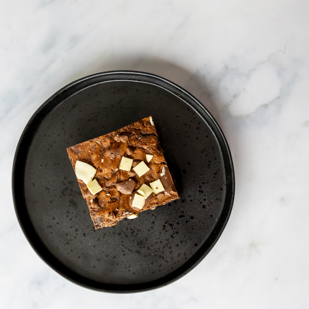 Square of brownies with white, milky, and black chocolate chips on a black plate against a marble background