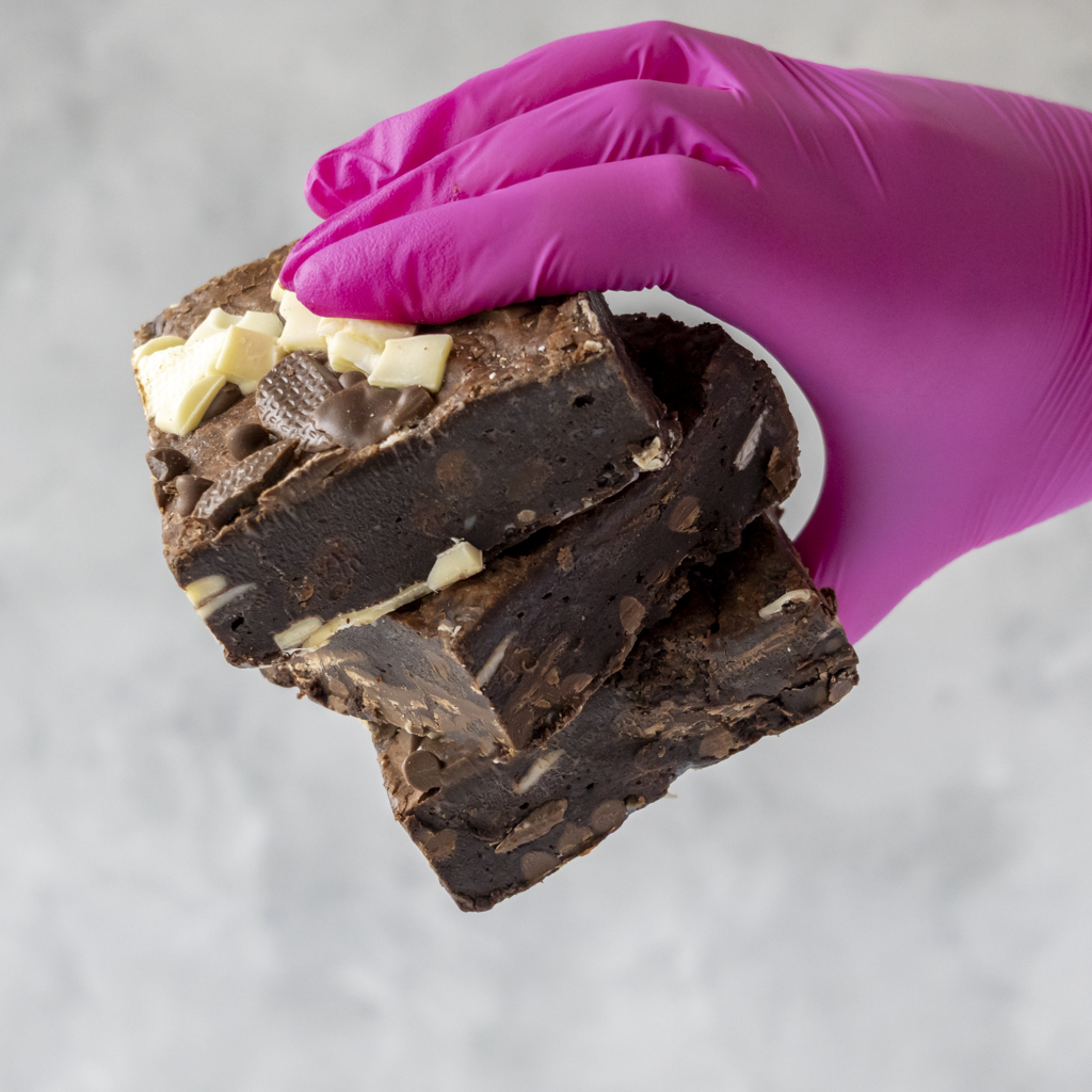 Hand wearing a pink glove holding a stack of Triple Chocolate Brownie Slab with different colour chocolate chips.