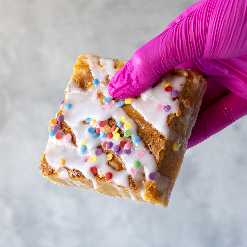 Birthday Blondie Slab with white chocolate and colorful sprinkles held by a hand in a pink glove, perfect for serving birthday parties or celebration occasions
