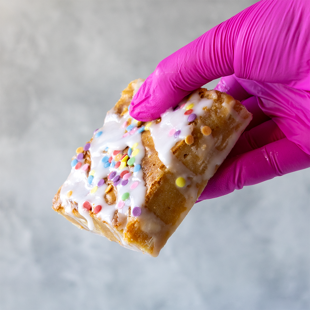 Hand wearing a pink glove holding a Birthday Blondie Slab with white icing and colorful sprinkles against a light background