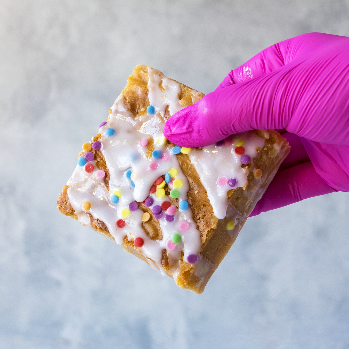 Hand holding a DoughGirl Birthday Blondie Slab with white chocolate topping and colourful sprinkles , wearing a pink glove on a light gray background.