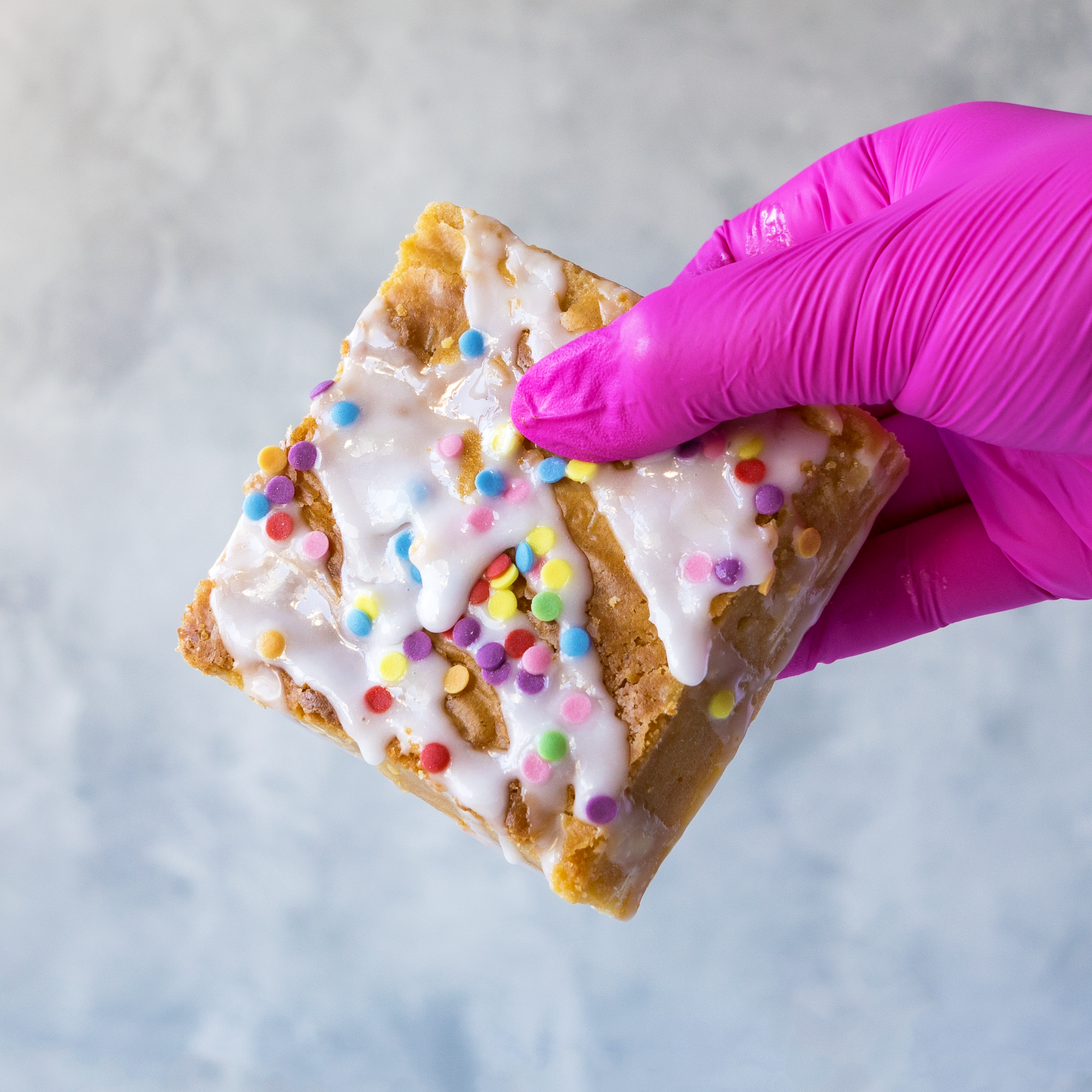 Hand holding a DoughGirl Birthday Blondie Slab with white chocolate topping and colourful sprinkles , wearing a pink glove on a light gray background.