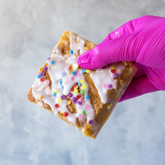 Hand holding a DoughGirl Birthday Blondie Slab with white chocolate topping and colourful sprinkles , wearing a pink glove on a light gray background.