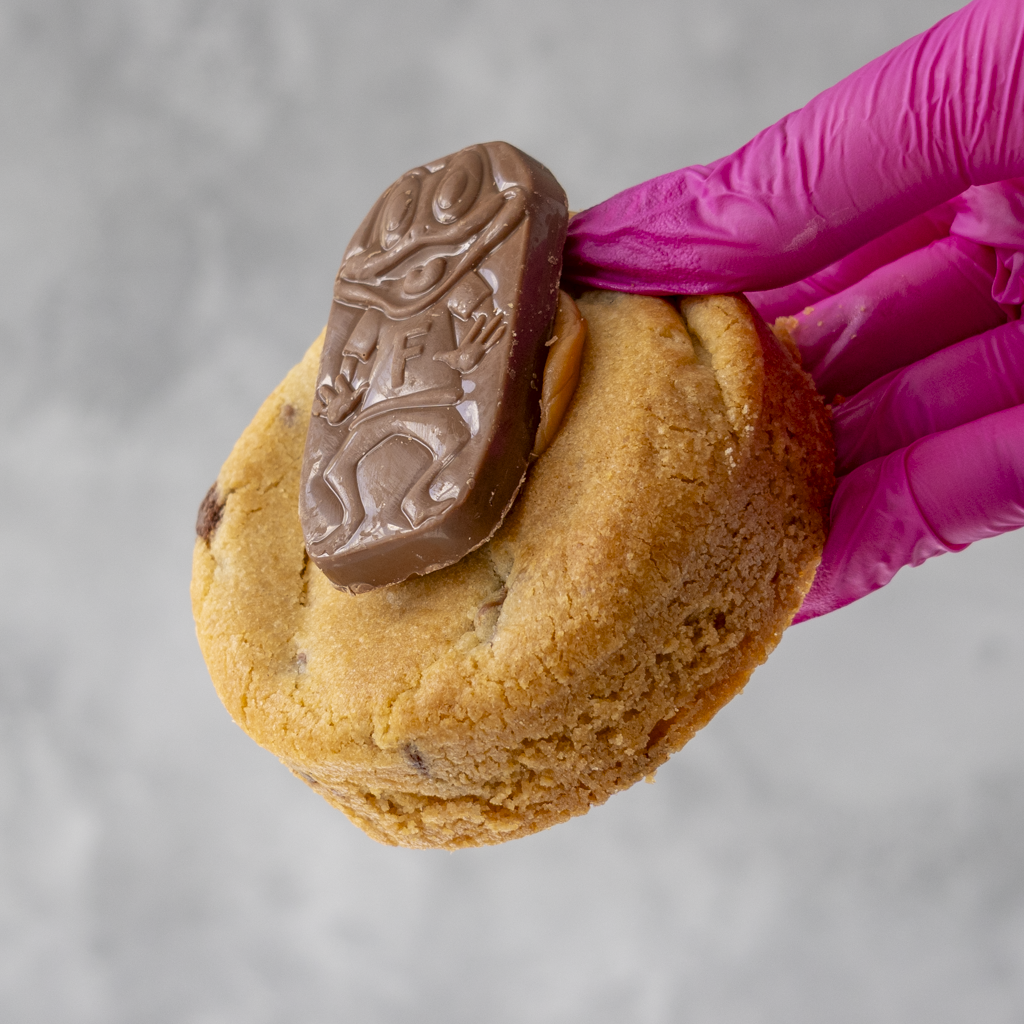 Caramel Freddo Deluxe Cookie with a Freddo chocolate held by a pink-gloved hand against a gray background