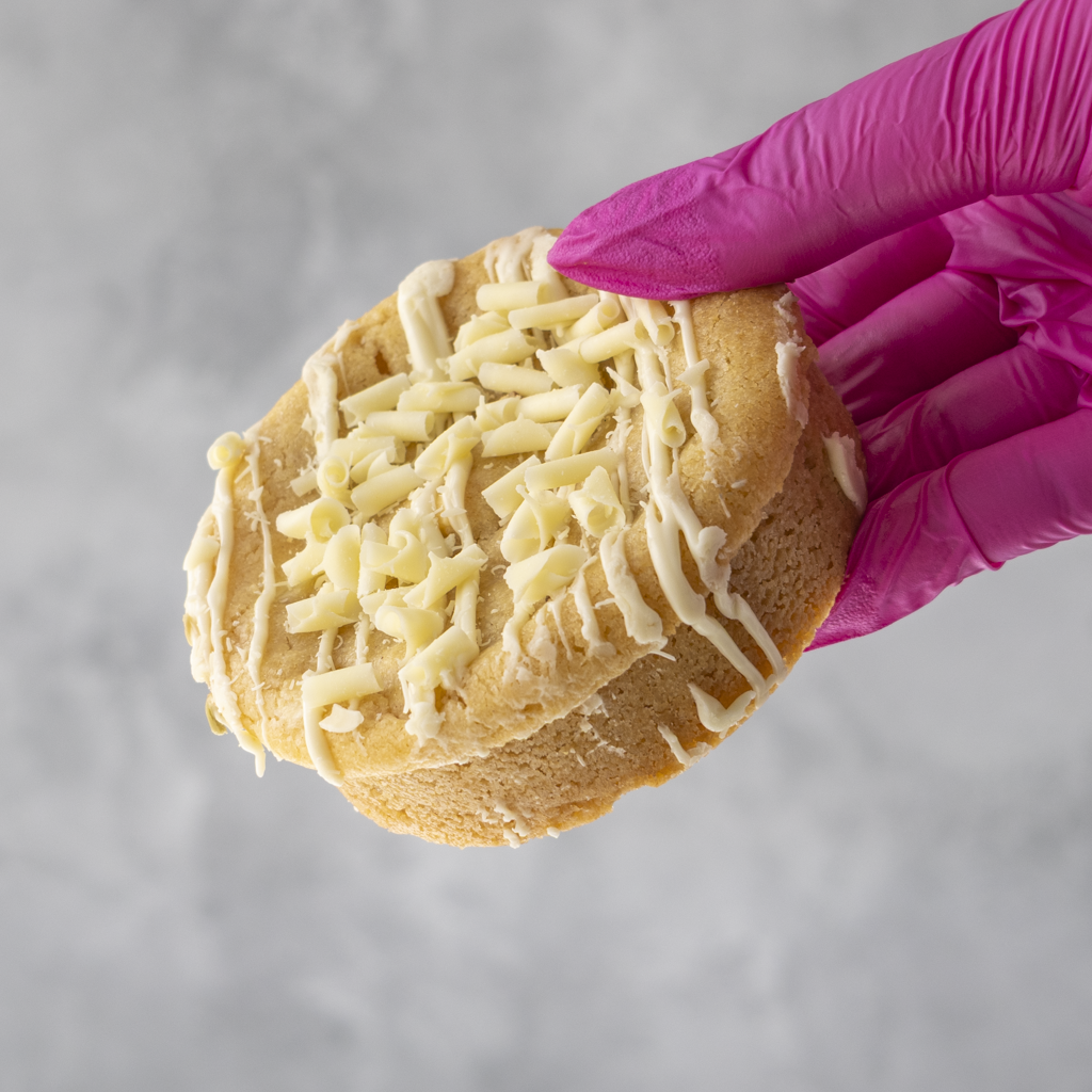 Lemon and White Chocolate Stuffed Deluxe Cookie with white frosting and being held by a gloved hand against a gray background