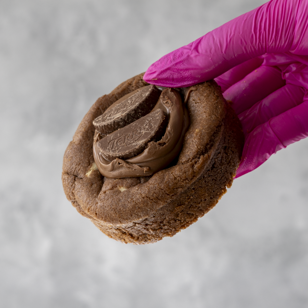 Chocolate Orange Deluxe Cookie with chocolate filling held by a pink-gloved hand on a gray background