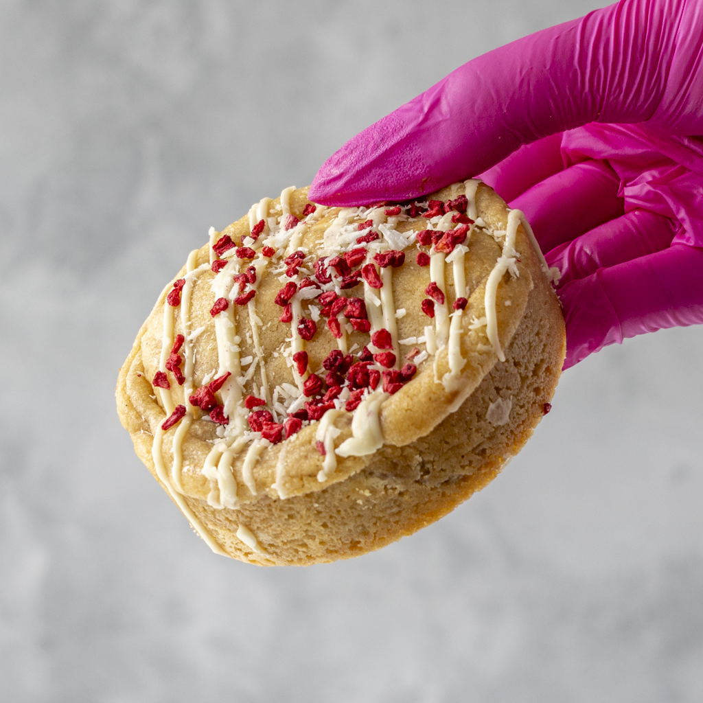 Raspberry, Coconut &amp; White Chocolate Deluxe Cookie with red and white toppings held by a hand in a pink glove on a gray background