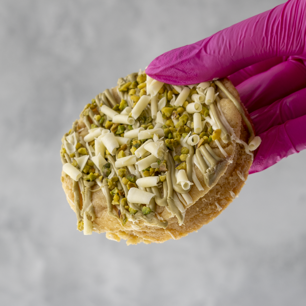Pistachio & White Chocolate Stuffed Deluxe Cookie with white and green icing held by a gloved hand against a gray background