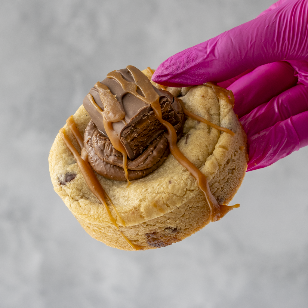 Mars Deluxe Cookie with chocolate filling and caramel drizzle held by a hand wearing a pink glove on a gray background