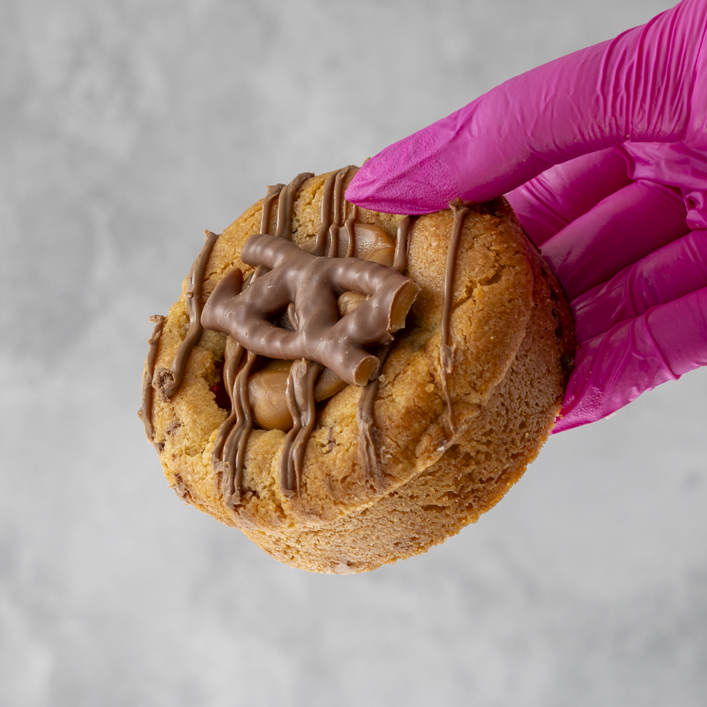 Curly Wurly Deluxe Cookie with chocolate drizzle held by a hand wearing a pink glove on a gray background