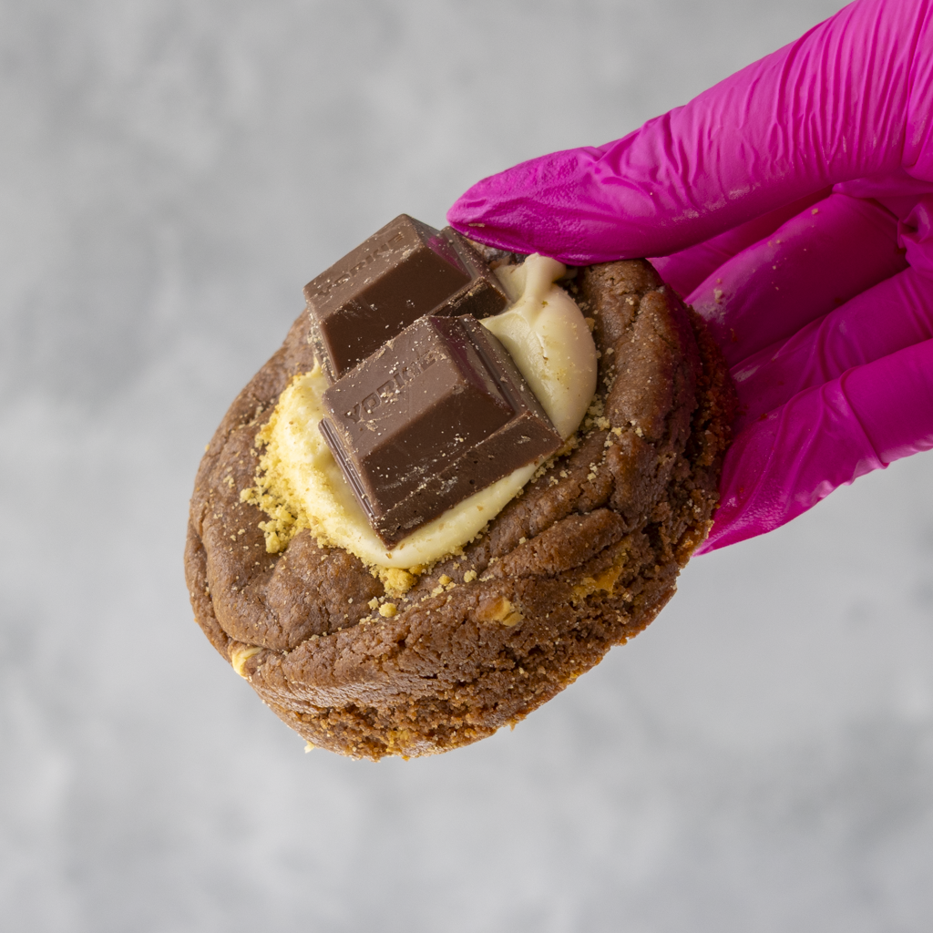 Yorkie, Biscuit & Brownie Deluxe Cookie with chocolate and cream filling held by a hand wearing a pink glove on a gray background