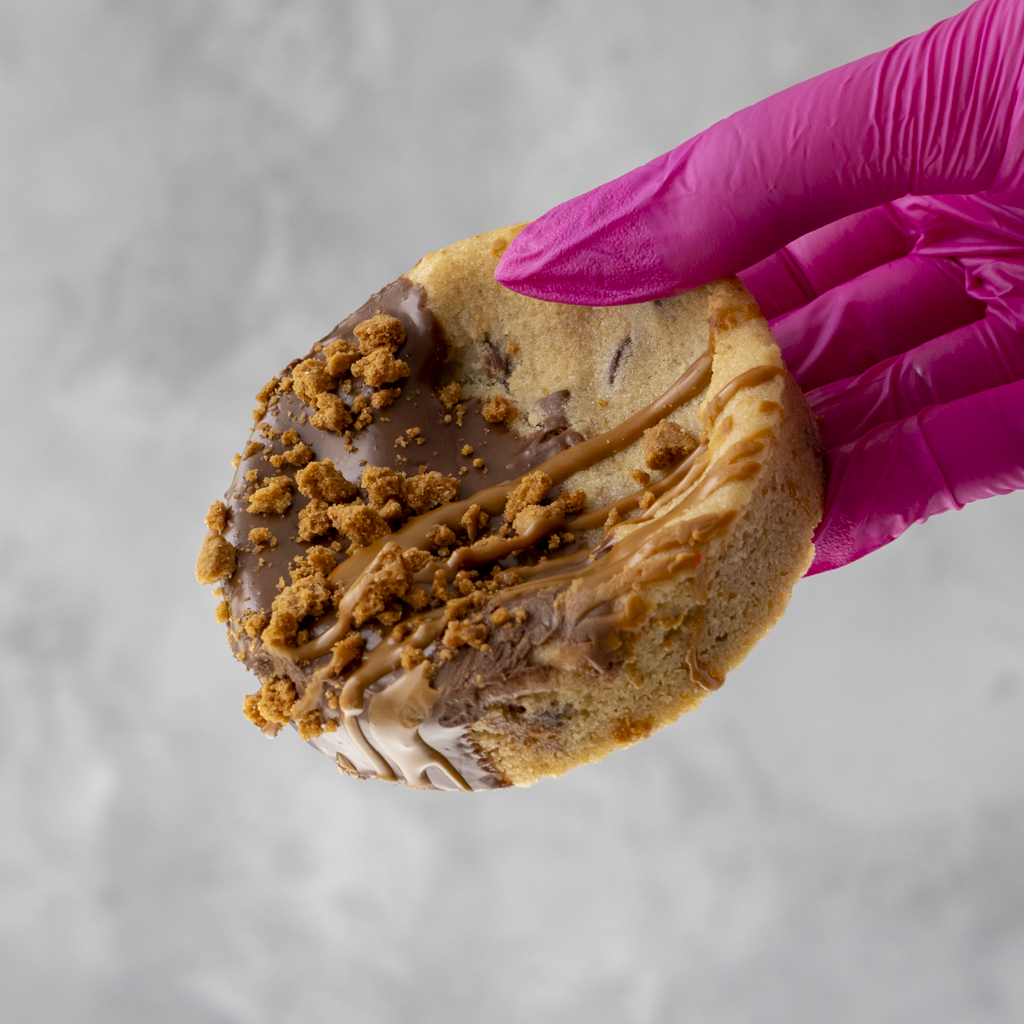 Hand wearing a pink glove holding a Biscella Deluxe Cookie with chocolate and crumb toppings against a gray background