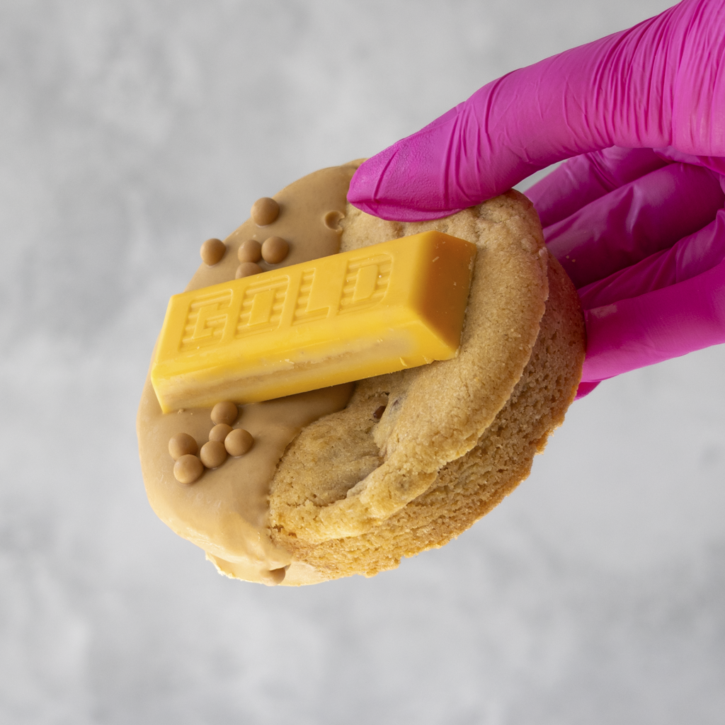 Hand wearing a pink glove holding a cookie with a gold chocolate bar on a gray background