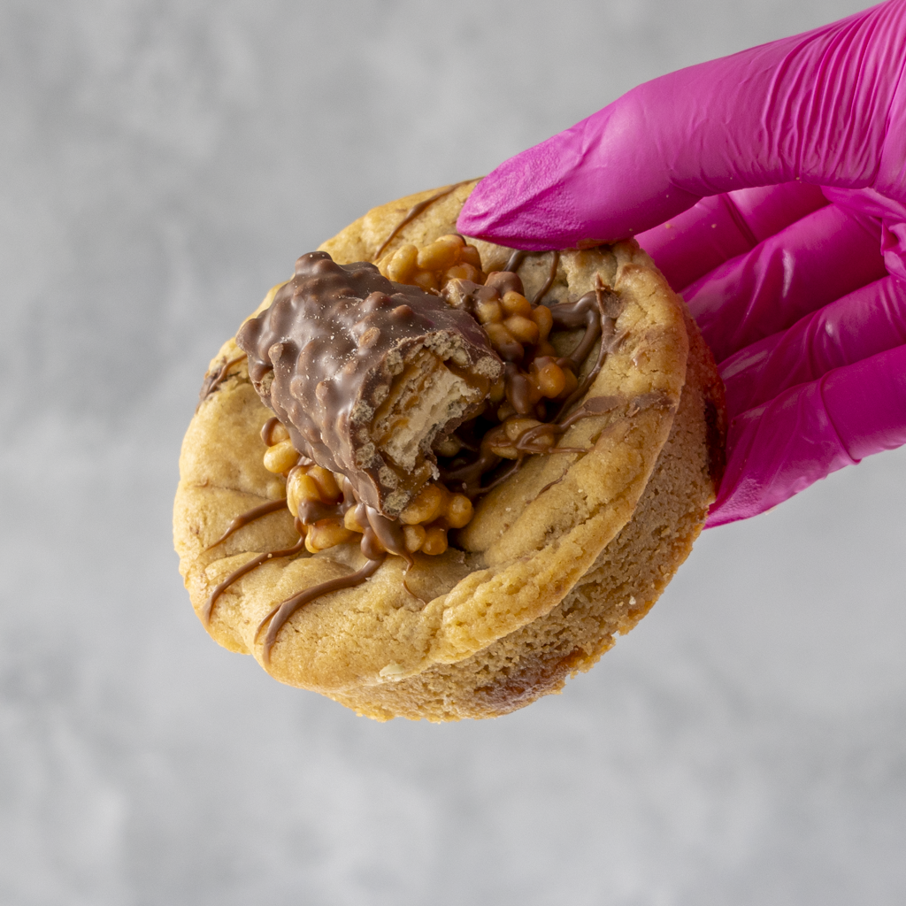 Lion Bar Stuffed Deluxe Cookie with chocolate filling held by a gloved hand against a gray background