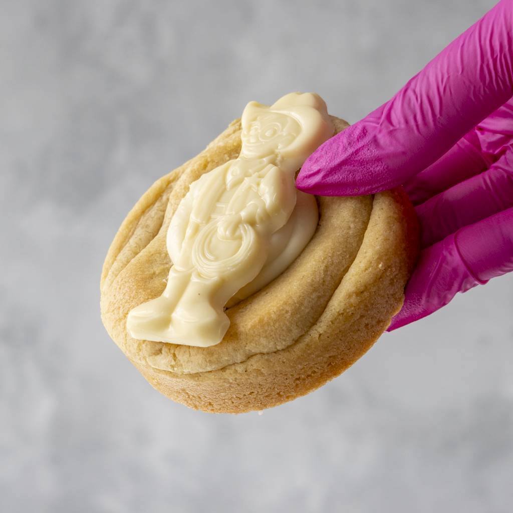Milkybar Bliss Deluxe Cookie with white icing shaped like a cat, held by a hand wearing a pink glove on a gray background