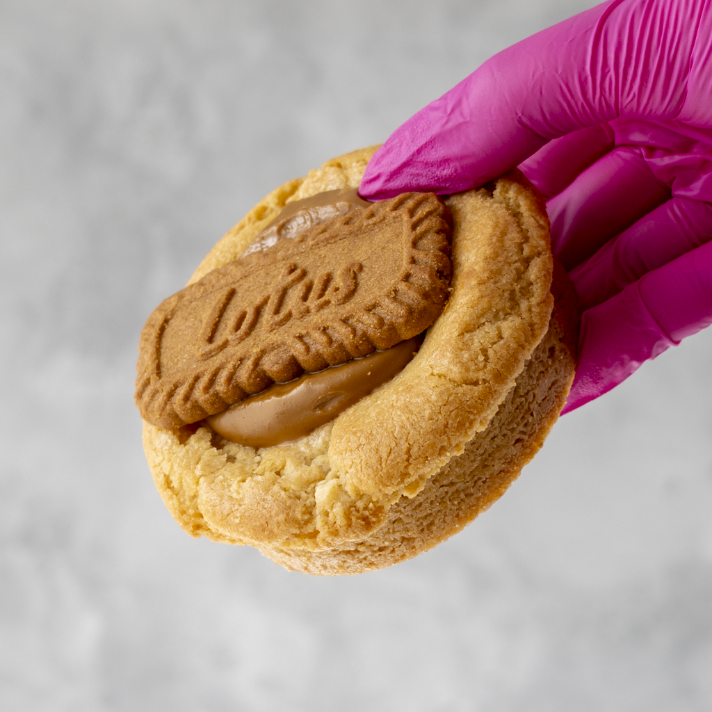 Hand wearing a pink glove holding a Biscoff Deluxe Cookie with a visible lotus biscoff against a gray background