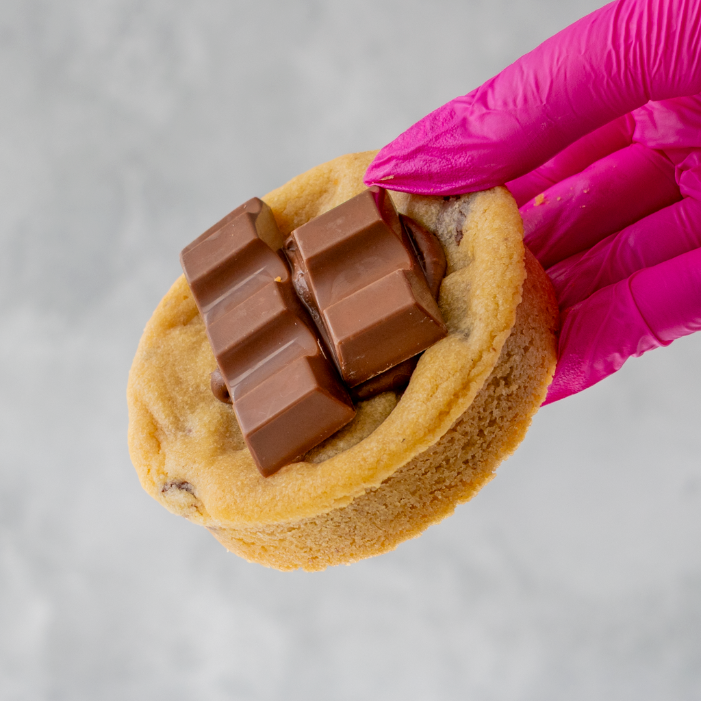 Hand wearing a pink glove holding a Kindertella Deluxe Cookie with chocolate bars on a gray background
