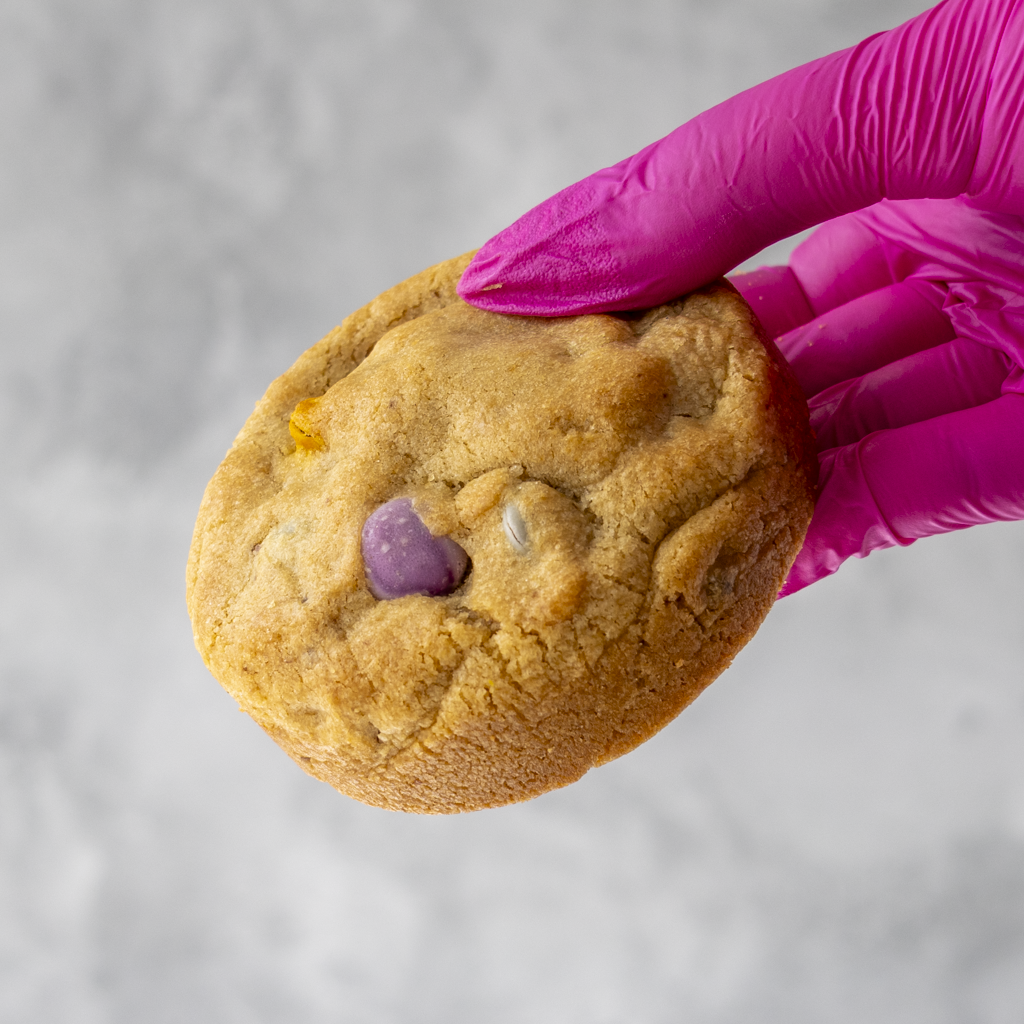 Rainbow Deluxe Cookie with a purple candy piece held by a pink gloved hand on a neutral background