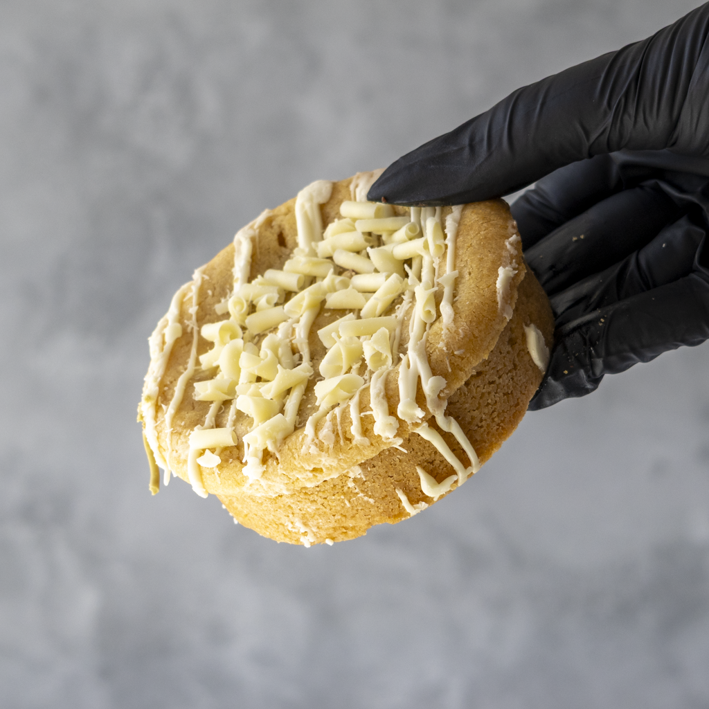 Lemon deluxe cookie with white chocolate shavings held by a black gloved hand against a gray background