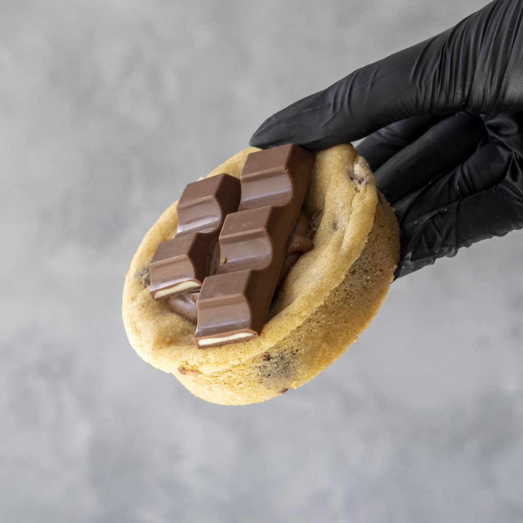 Hand wearing a black glove holding a Kindertella Deluxe Cookie with chocolate bars on a gray background