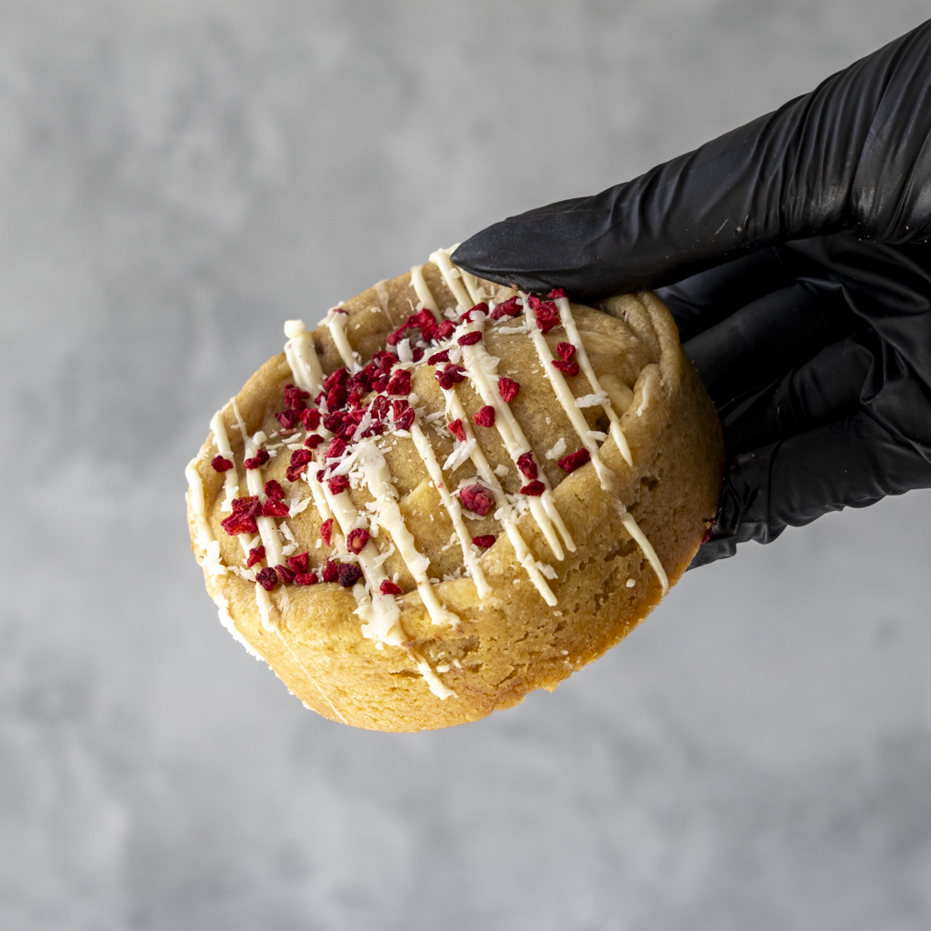 DoughGirl Raspberry, Coconut &amp; White Chocolate Deluxe Cookie with white drizzle and red toppings held by a black gloved hand against a gray background
