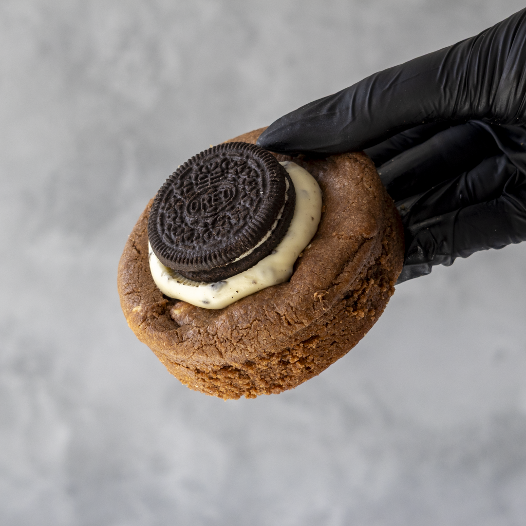 Hand wearing a black glove holding a DoughGirl Deluxe Cookie with an Oreo filling against a gray background