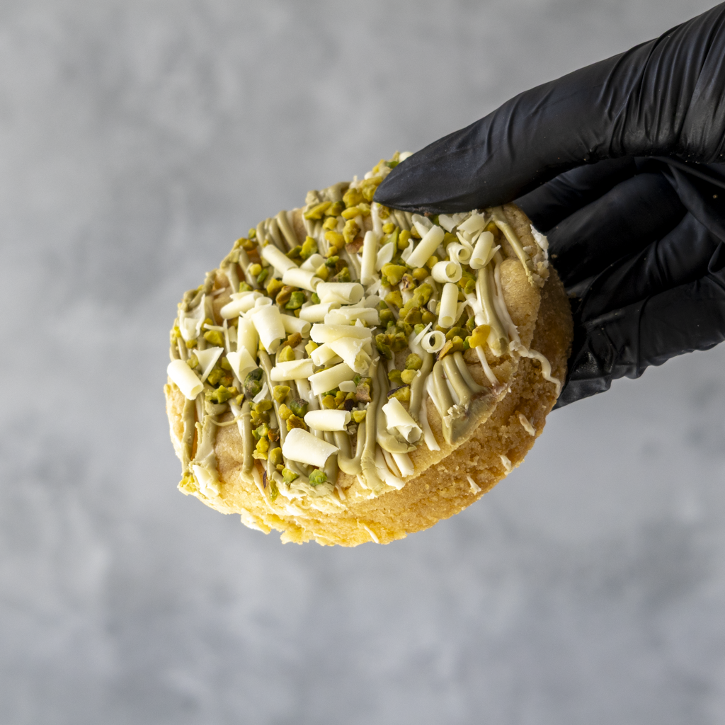 Hand holding a Pistachio & White Chocolate Stuffed Deluxe Cookie by DoughGirl UK with a black glove against a gray background