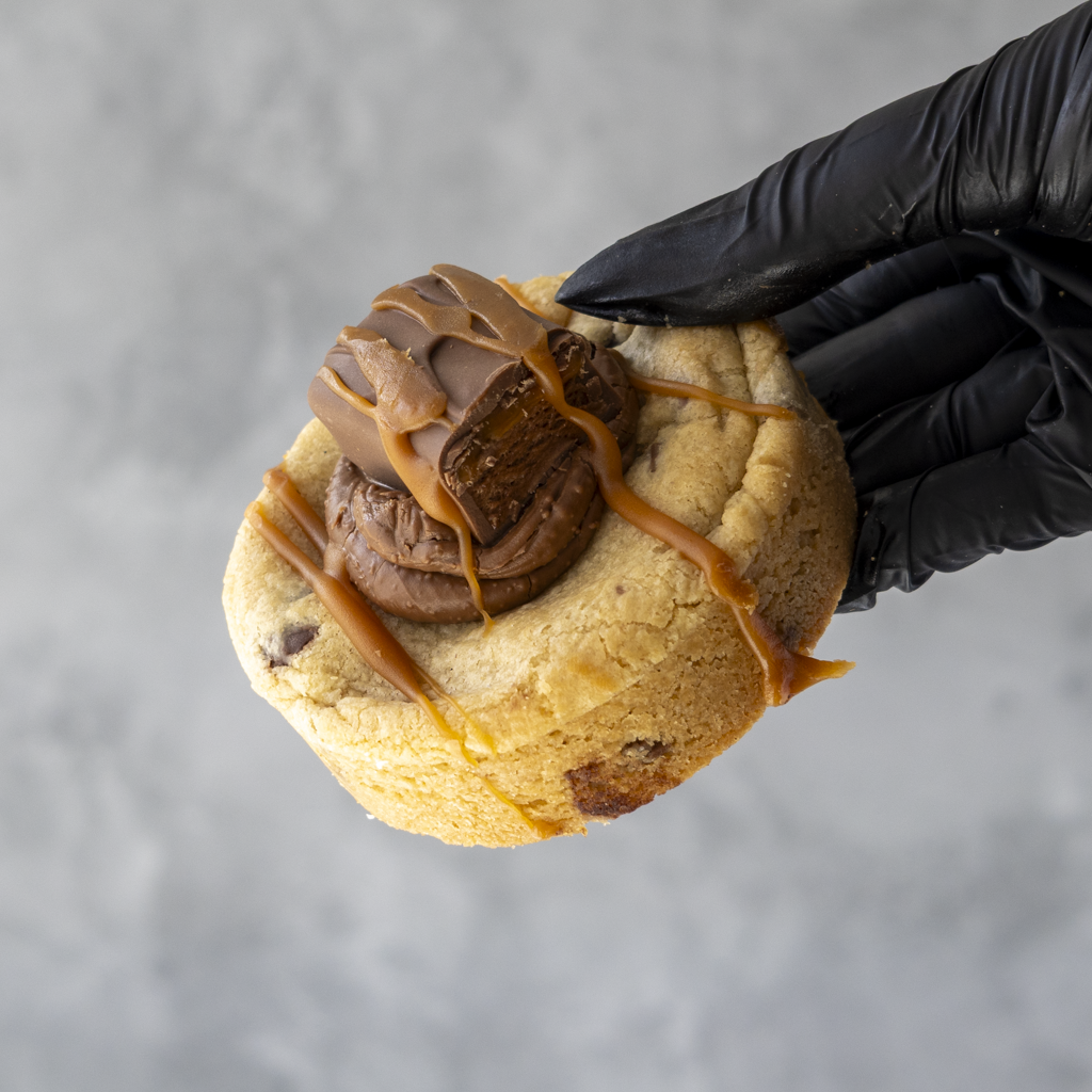 Cookie with Mars chocolate filling and caramel drizzle held by a black gloved hand against a gray background