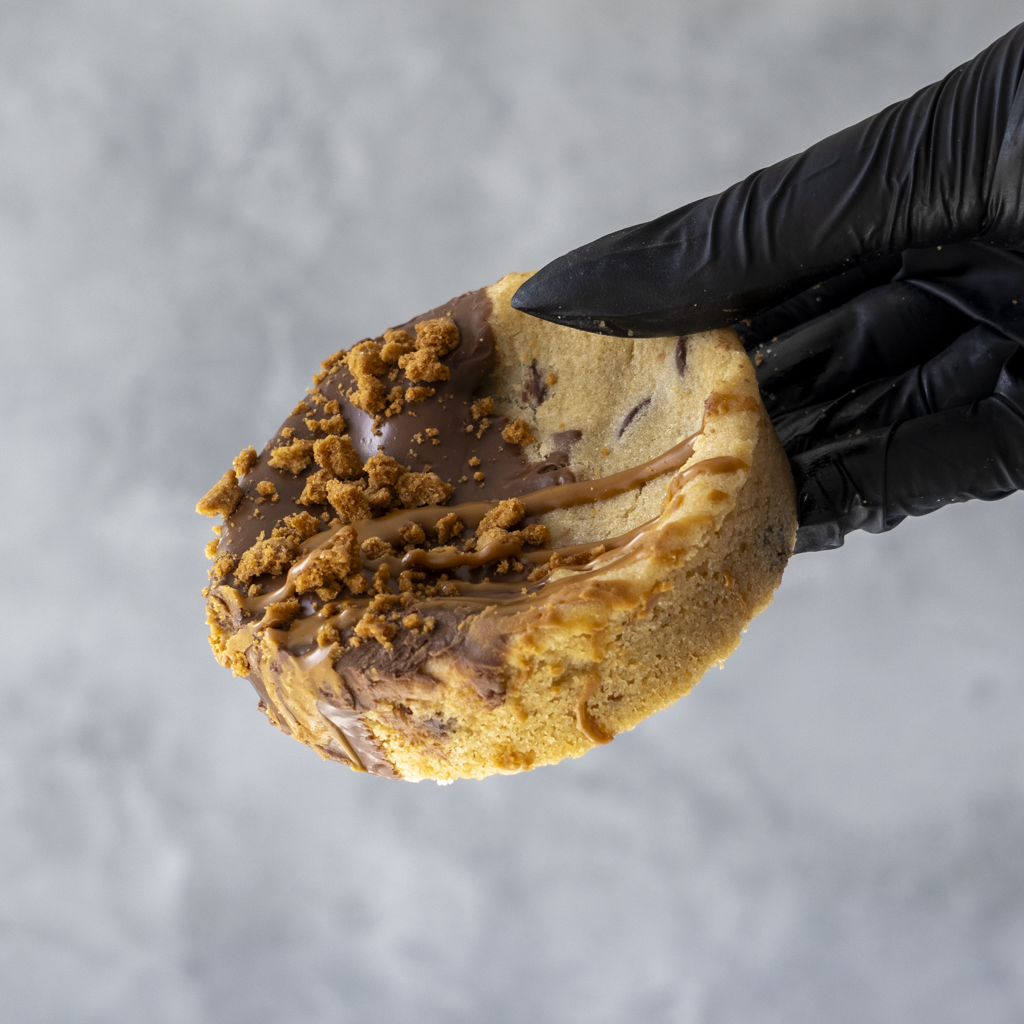 Black gloved hand holding a Biscella Deluxe Cookie with chocolate and crumb toppings against a gray background
