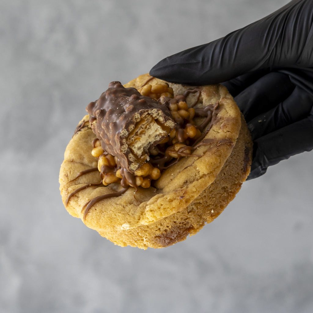 Hand holding a DoughGirl Lion Bar Stuffed Deluxe Cookie with chocolate bar and nuts against a gray background