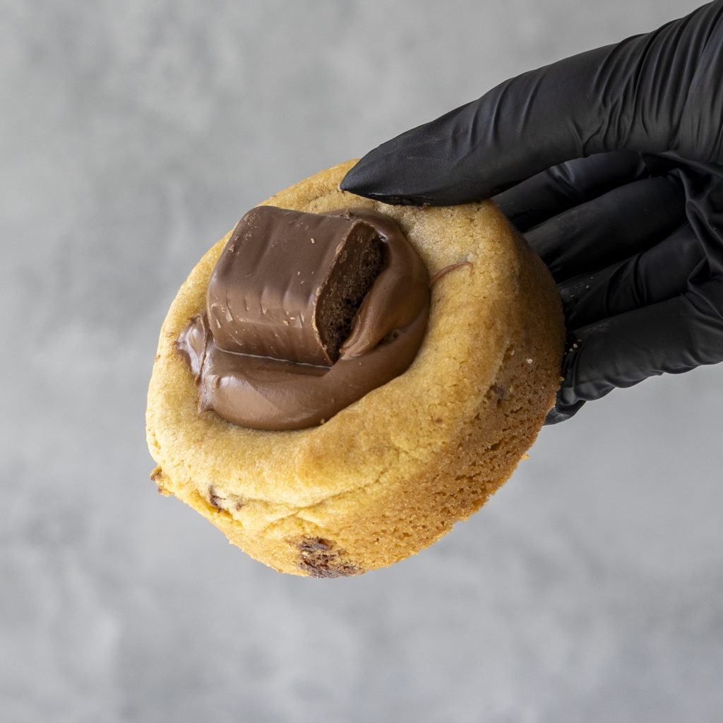 Hand wearing a black glove holding a DoughGirl Cadbury Wispa Deluxe Cookie with cadbury chocolate against a gray background