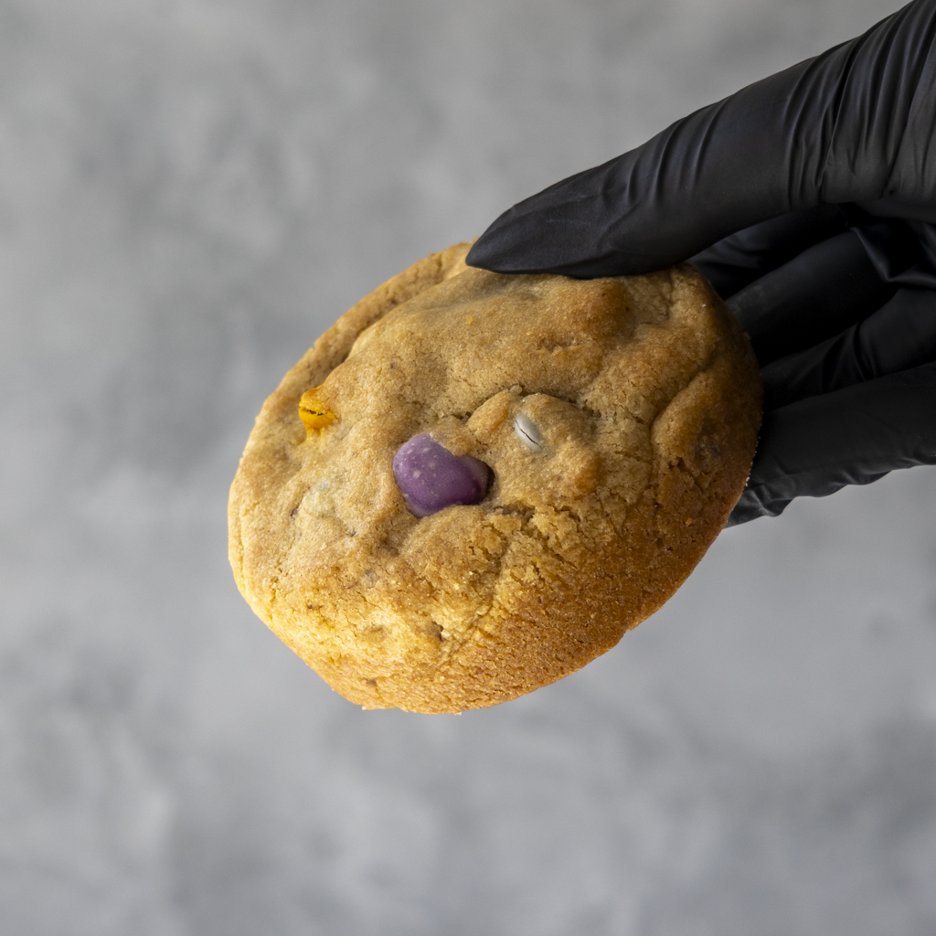 A colorful Rainbow Deluxe Cookie by DoughGirl held by a black gloved hand against a gray background