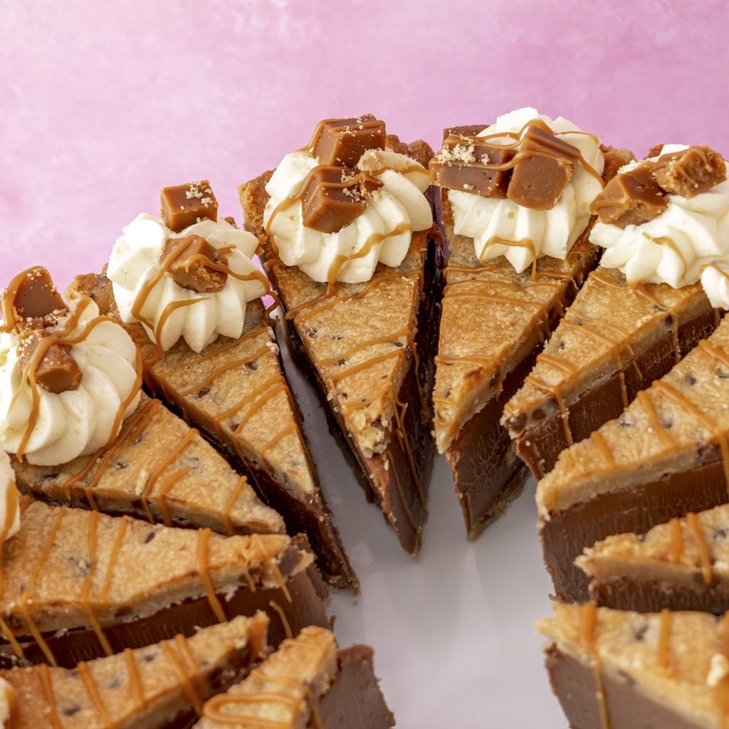 Full sliced Banoffee Cookie Pie by DoughGirl UK with whipped cream and caramel drizzle on a pink background