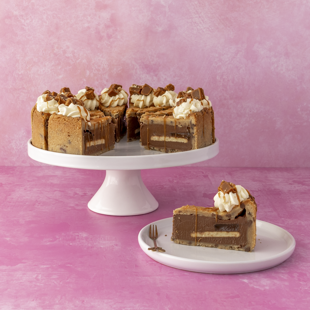 Full 12-Slice Banoffee Cookie Pie by DoughGirl UK with white frosting on a white cake stand against a pink background