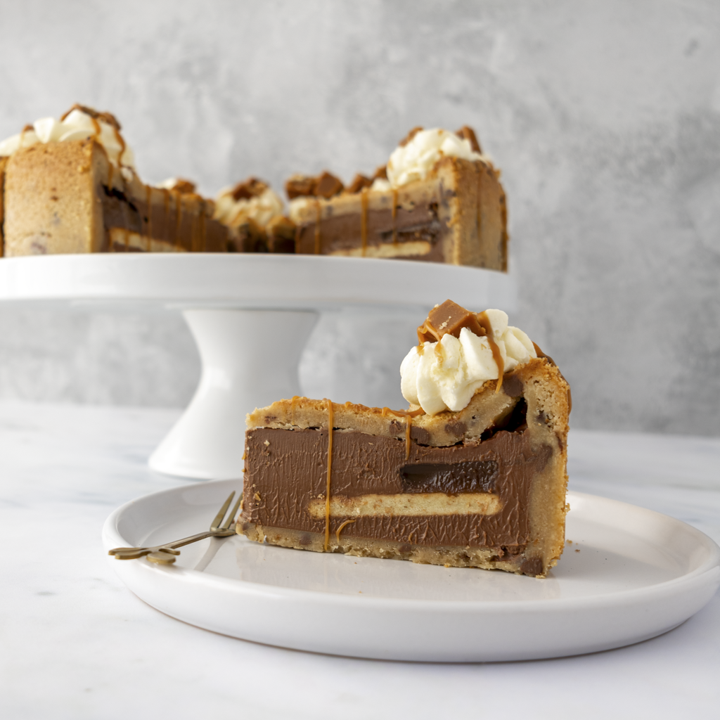 Slice of layered chocolate Banoffee Cookie Pie topped with white cream on a white plate, with the rest of the cake in the background.