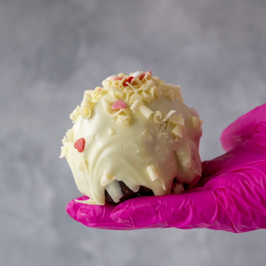 Hand holding a DoughGirl Filthy Fluff Brookie Choccy Boom Bites with red heart sprinkles against a gray background