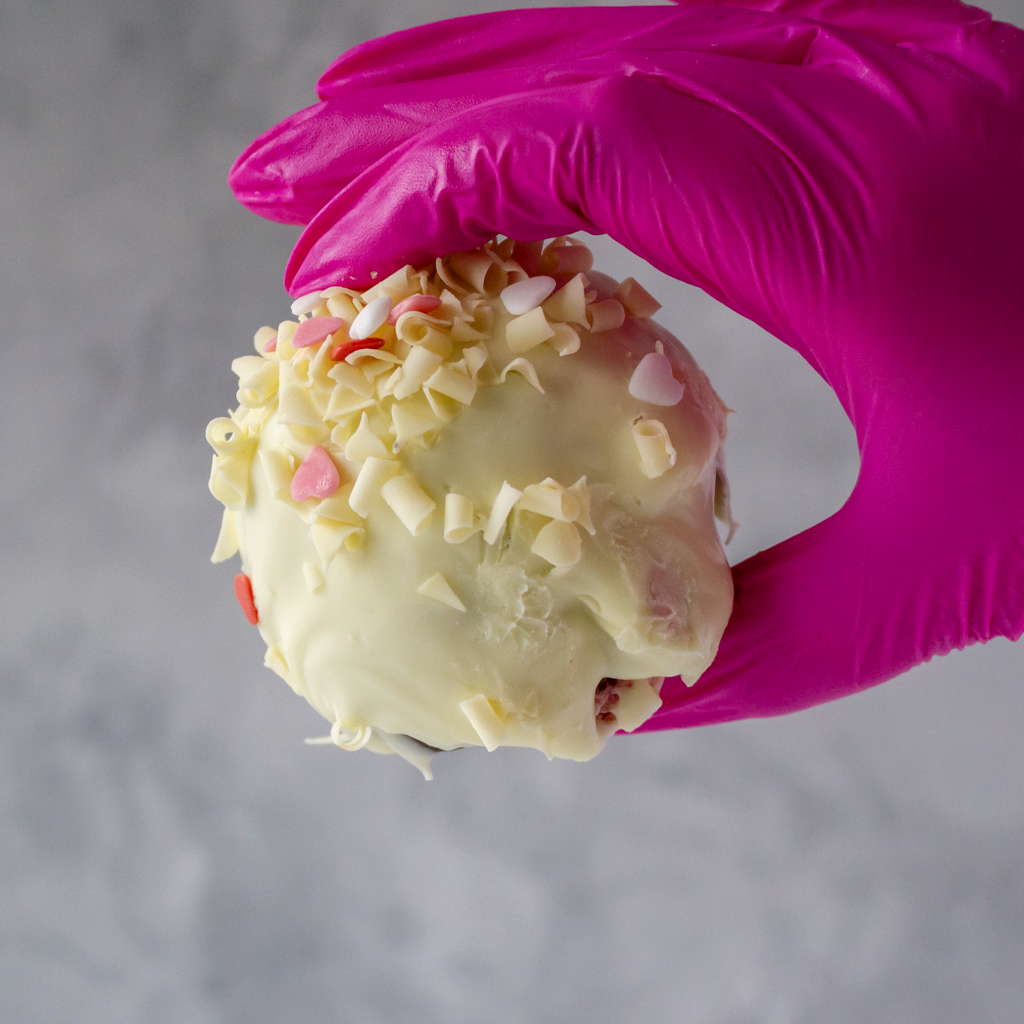 Filthy Fluff Brookie Choccy Boom Bites by DoughGirl with white chocolate and pink heart-shaped sprinkles held by a gloved hand against a gray background
