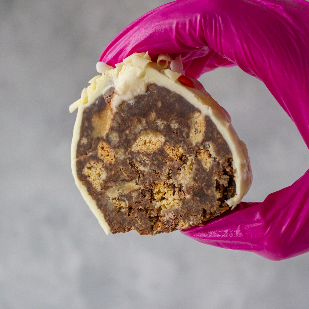 Half-eaten Filthy Fluff Brookie Choccy Boom Bites with classic milk chocolate chip cookie, rich chocolate brownie and gooey marshmallow swirl, held by a gloved hand against a neutral background