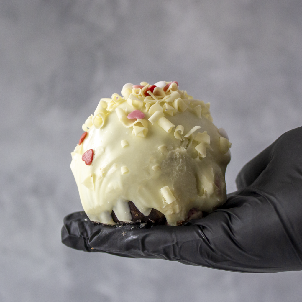 Hand holding a Filthy Fluff Brookie Choccy Boom Bites by DoughGirl UK with white chocolate and red candies against a gray background