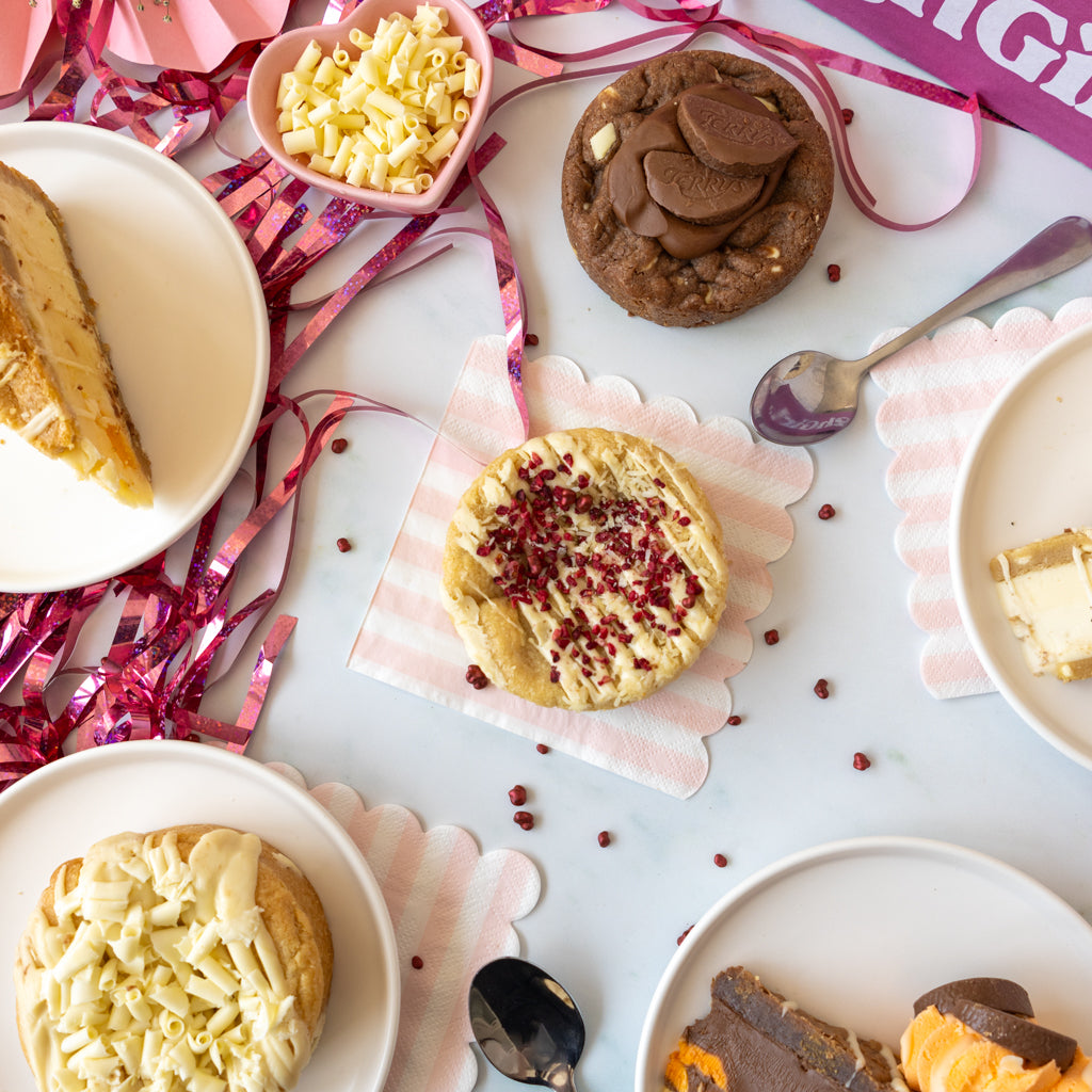 An overhead styled flat-lay of assorted baked desserts on white plates and a napkin, surrounded by pink and red tinsel ribbons. Visible items include a chocolate chip cookie, a chocolate cookie pie, a cookie with white chocolate chips, and a slice of cookie pie on a plate.