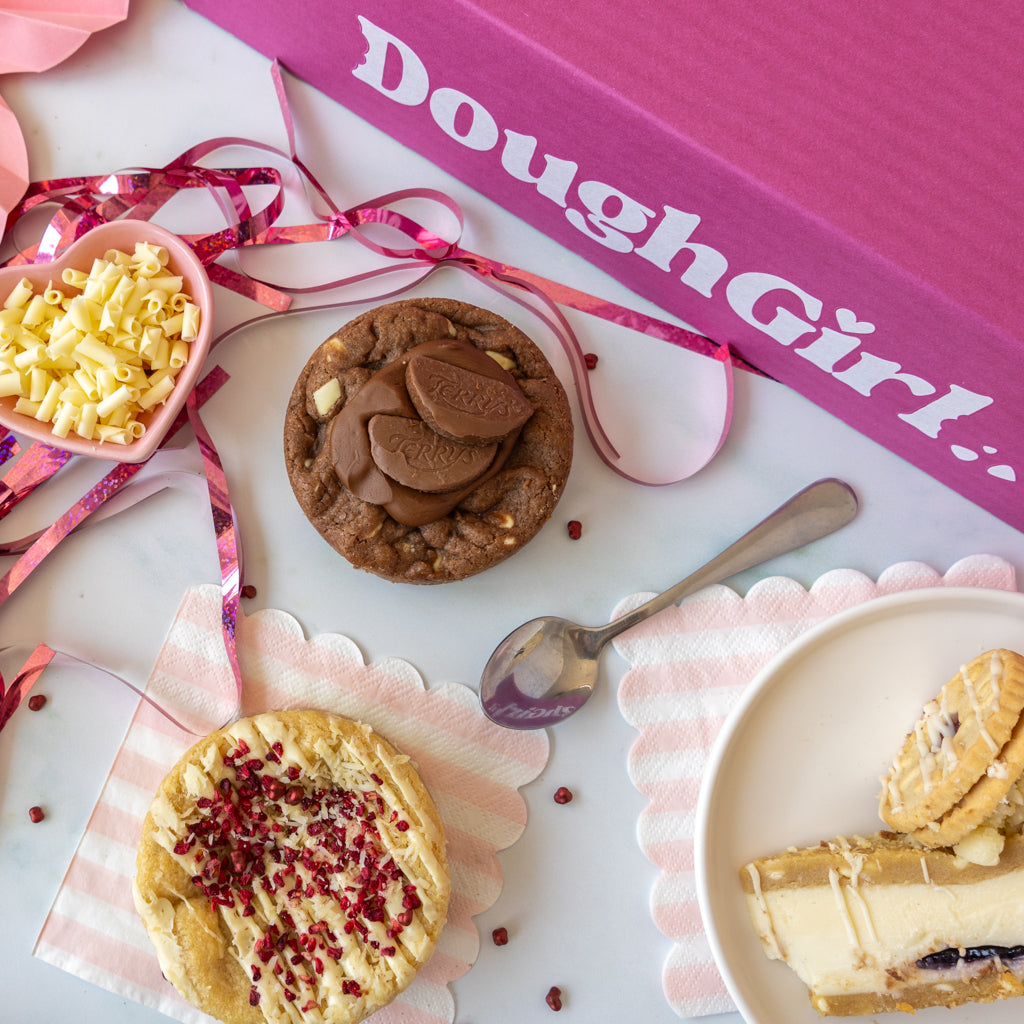 An overhead styled flat-lay of baked goods on a white surface with pink accents. A pink box labeled 'DoughGirl.