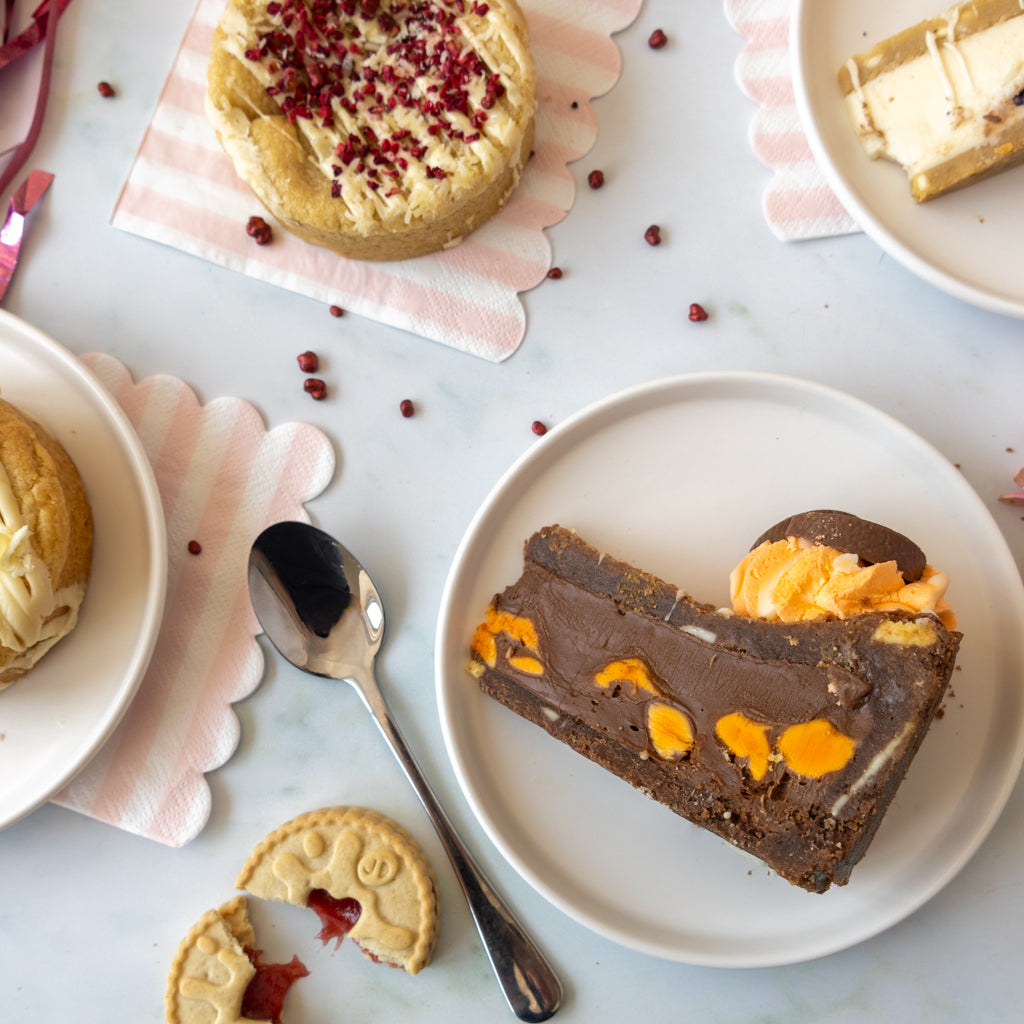 A styled overhead flat-lay of assorted baked goods on white plates and pink striped napkins. The main focus is a slice of chocolate cake with orange fruit pieces on a white plate, alongside other pastries including a tart with red berry topping and cream-filled pastry.