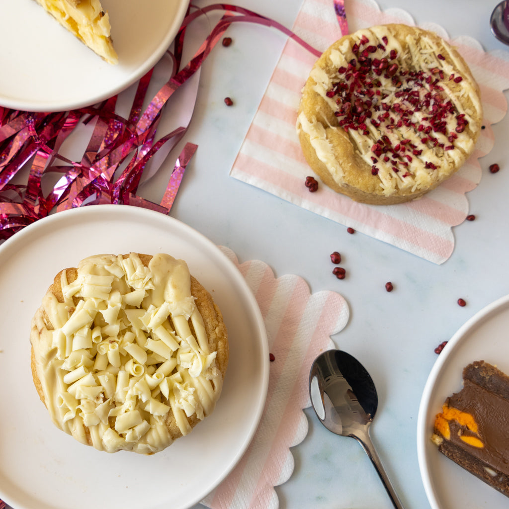 Two decorated cookies on white plates on a light marble surface, with pink ribbon, a spoon, and partial dessert slices in a styled tabletop setting.