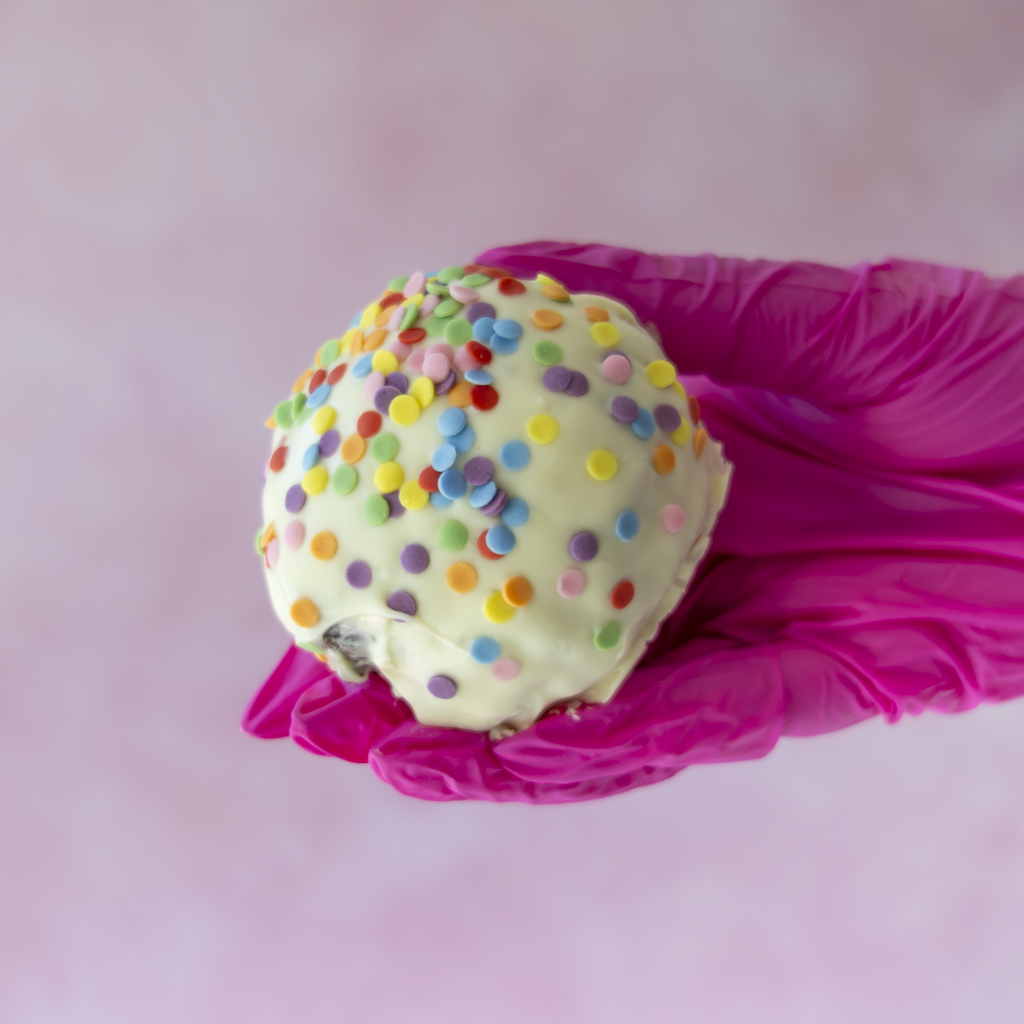 A white chocolate-covered Brookie Choccy Boom Bite decorated with colourful round sprinkles, held in a bright pink gloved hand against a soft pink background.