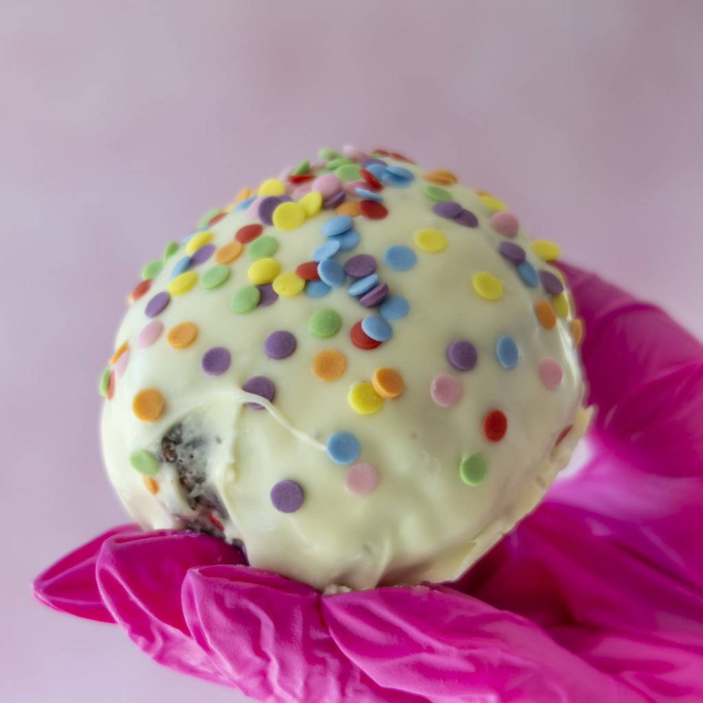 A close-up of a round white chocolate-covered Brookie Choccy Boom Bite topped with colourful round sprinkles, held in a bright pink gloved hand against a soft pink background.