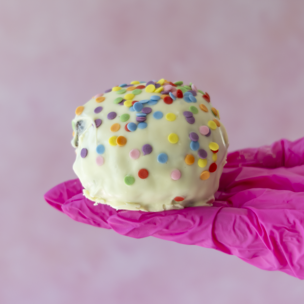 A spherical white chocolate-covered Brookie Choccy Boom Bite in colourful round sprinkles, held on a bright pink gloved hand against a soft pink background.